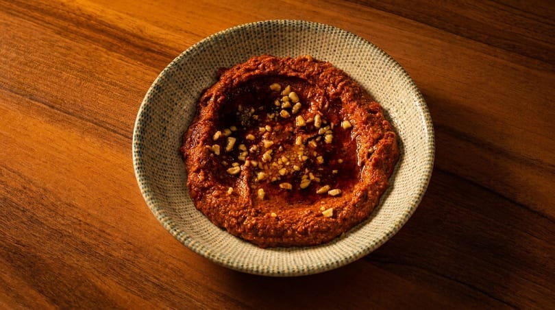 A rustic bowl of red muhammara dip, drizzled with olive oil and topped with chopped nuts.