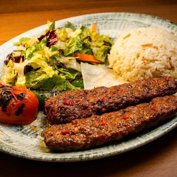 Two Adana kebabs with rice, salad, and a grilled tomato on a rustic plate.