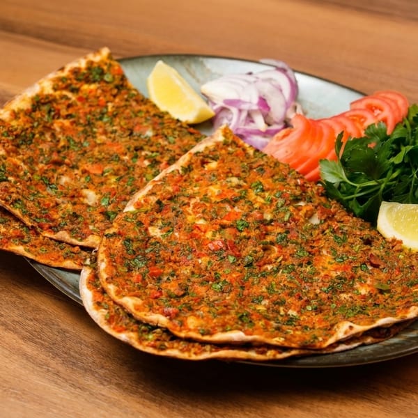 Three lahmacun flatbreads with minced meat and herbs, served with lemon, tomatoes, onions, and parsley.