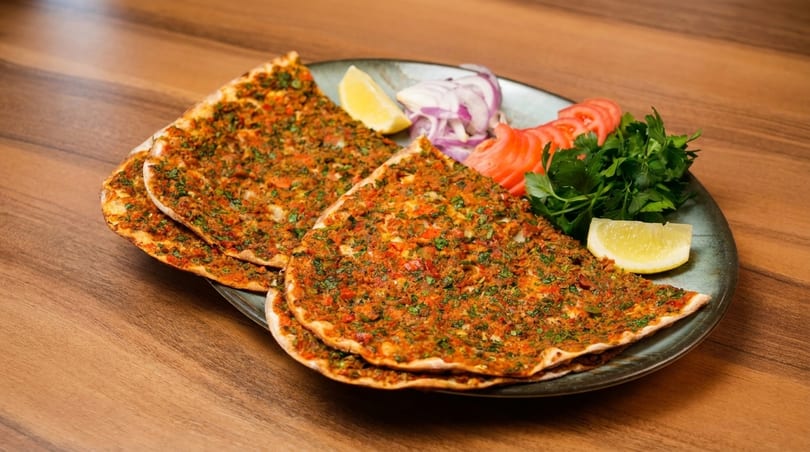 Three lahmacun flatbreads with minced meat and herbs, served with lemon, tomatoes, onions, and parsley.